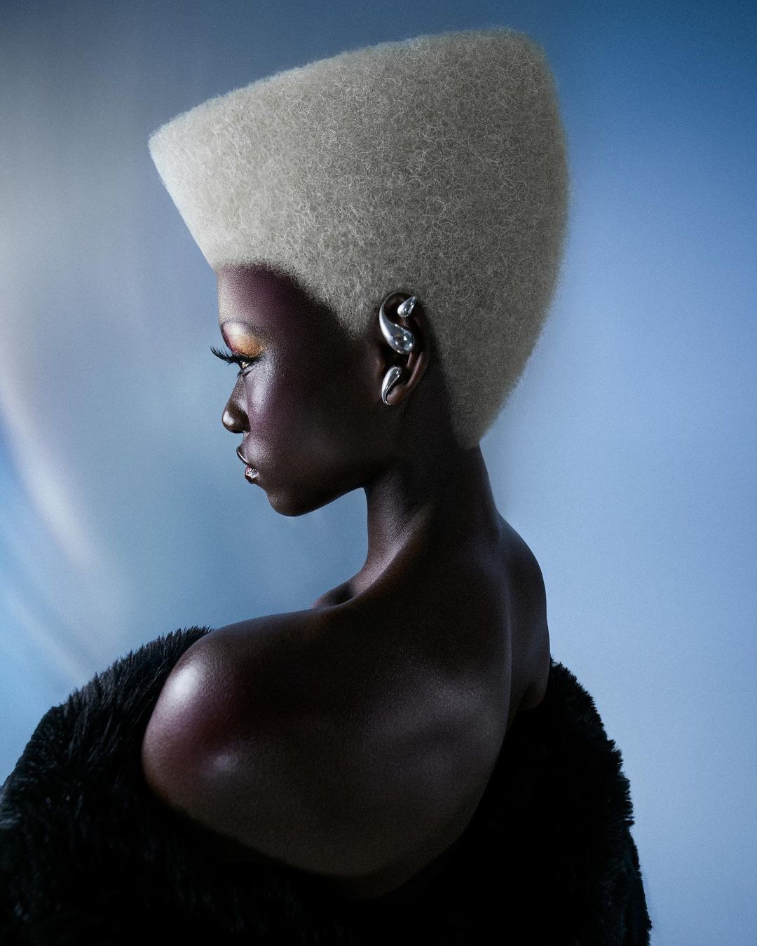 Fashion portrait of a Black female model with a boxy platinum blonde hairstyle, large earrings, and a black textured shawl on a blue gradient background.
