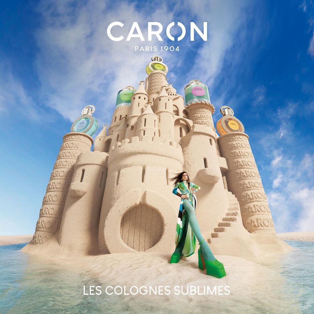 Caron fragrance campaign features a woman in green outfit next to a sandcastle with perfume bottles, under blue sky and ocean water.