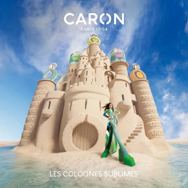 Caron fragrance campaign features a woman in green outfit next to a sandcastle with perfume bottles, under blue sky and ocean water.