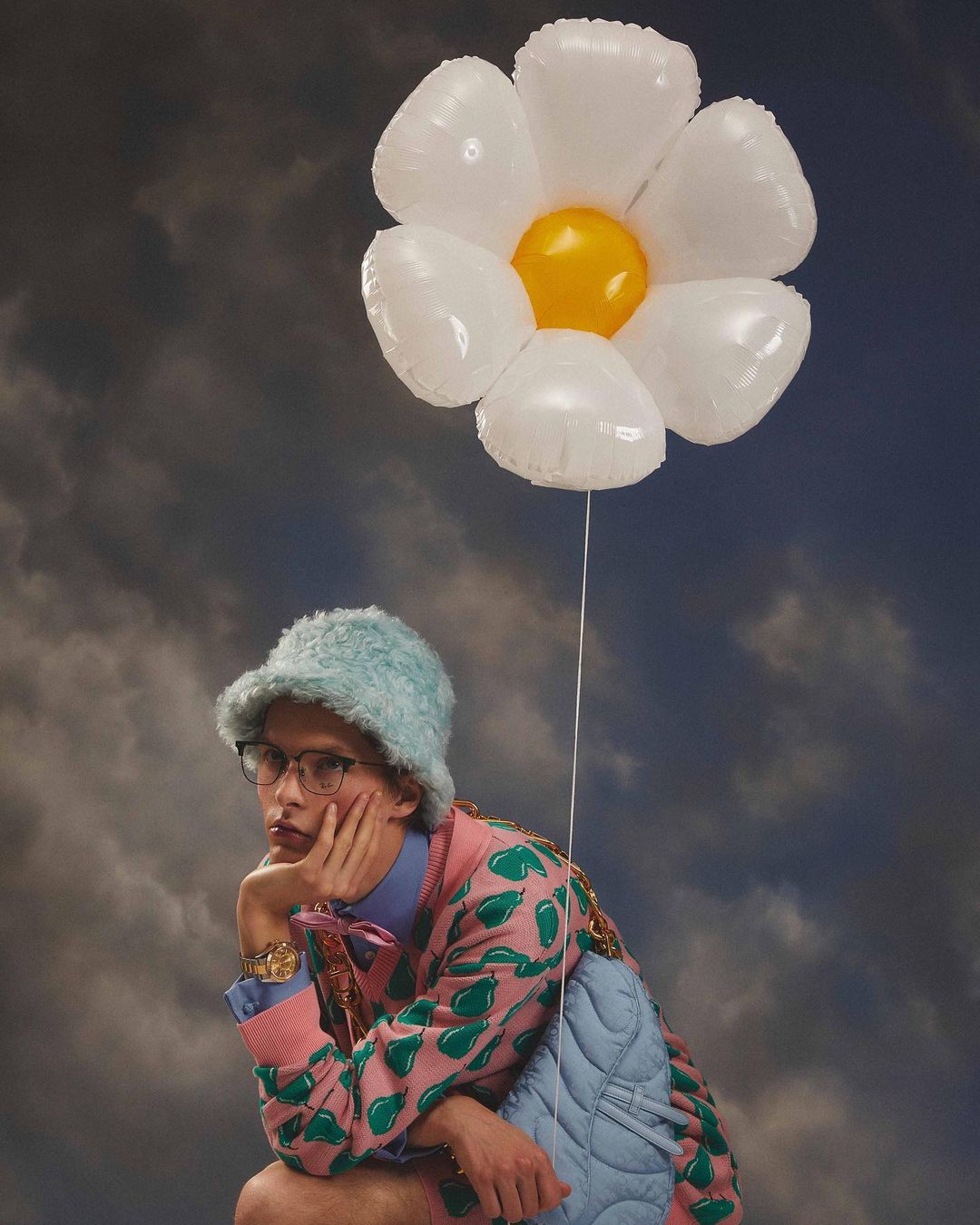 A contemplative adult male sits with a flower balloon against a cloudy backdrop, wearing glasses, a hat, and colorful patterned jacket.
