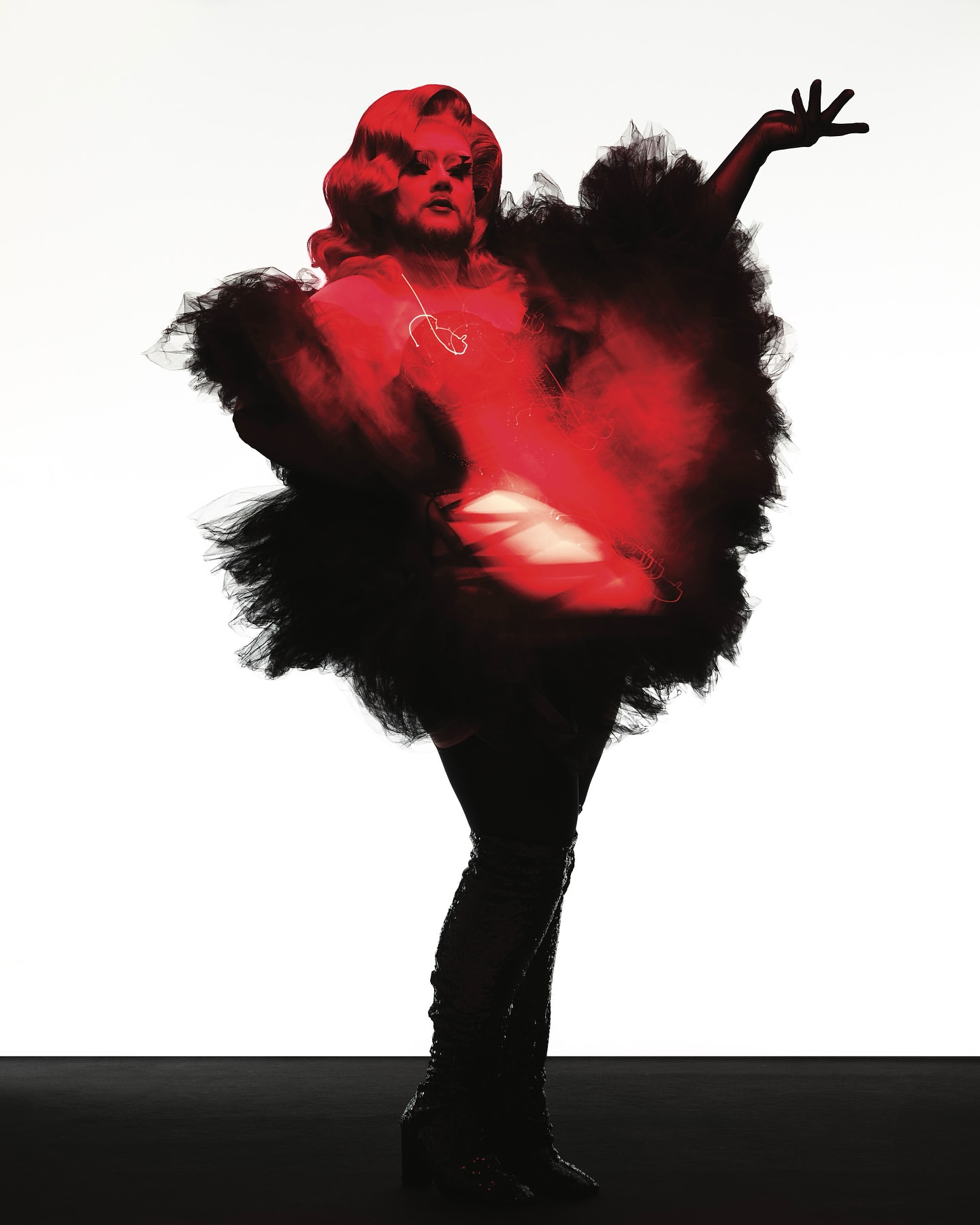Drag queen poses dramatically in studio wearing red and black feathered costume, red hair, and shiny black thigh-high boots.