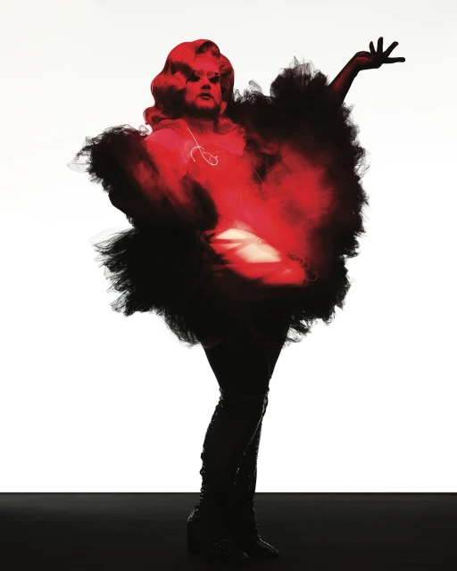 Drag queen poses dramatically in studio wearing red and black feathered costume, red hair, and shiny black thigh-high boots.