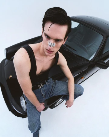 Stylish man poses with a black Porsche in a studio, wearing a black tank top, jeans, and a unique face accessory, captured in high angle.