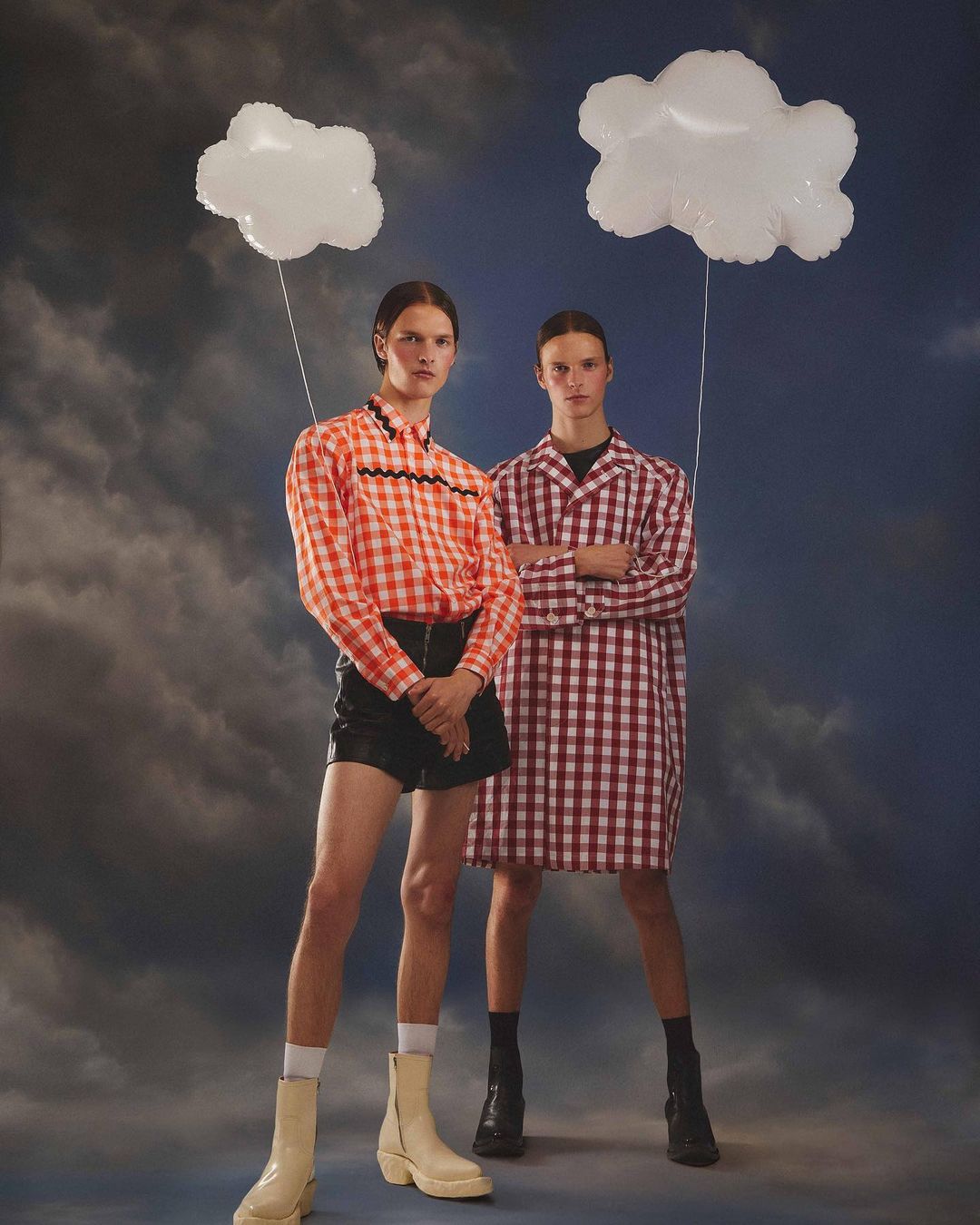 Two fashion models pose in gingham shirts and white boots with cloud balloons against a dreamy blue sky background. Studio lighting.
