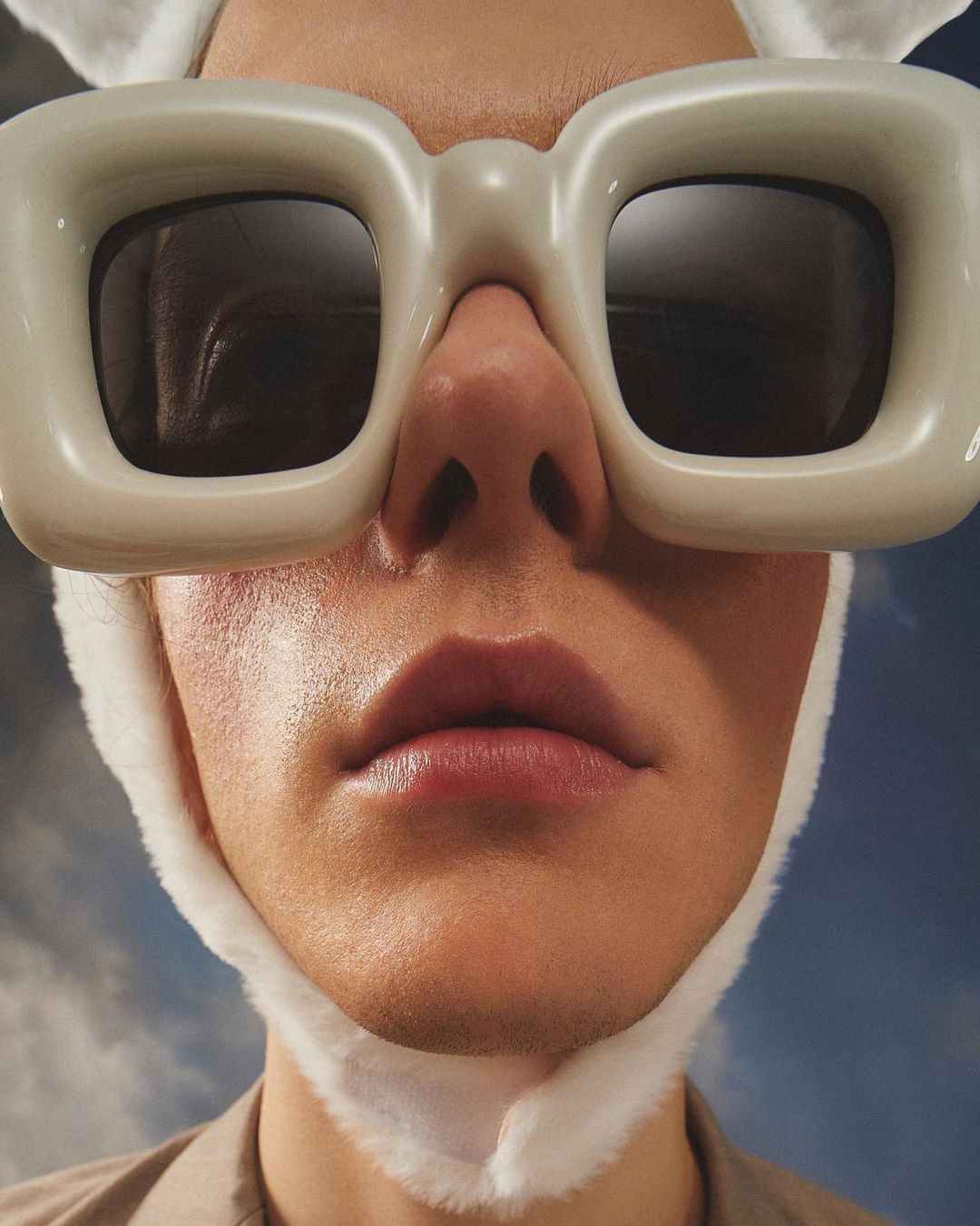 Close-up of a person wearing oversized sunglasses and bunny ears. Head and shoulders shot against a blurred sky background.