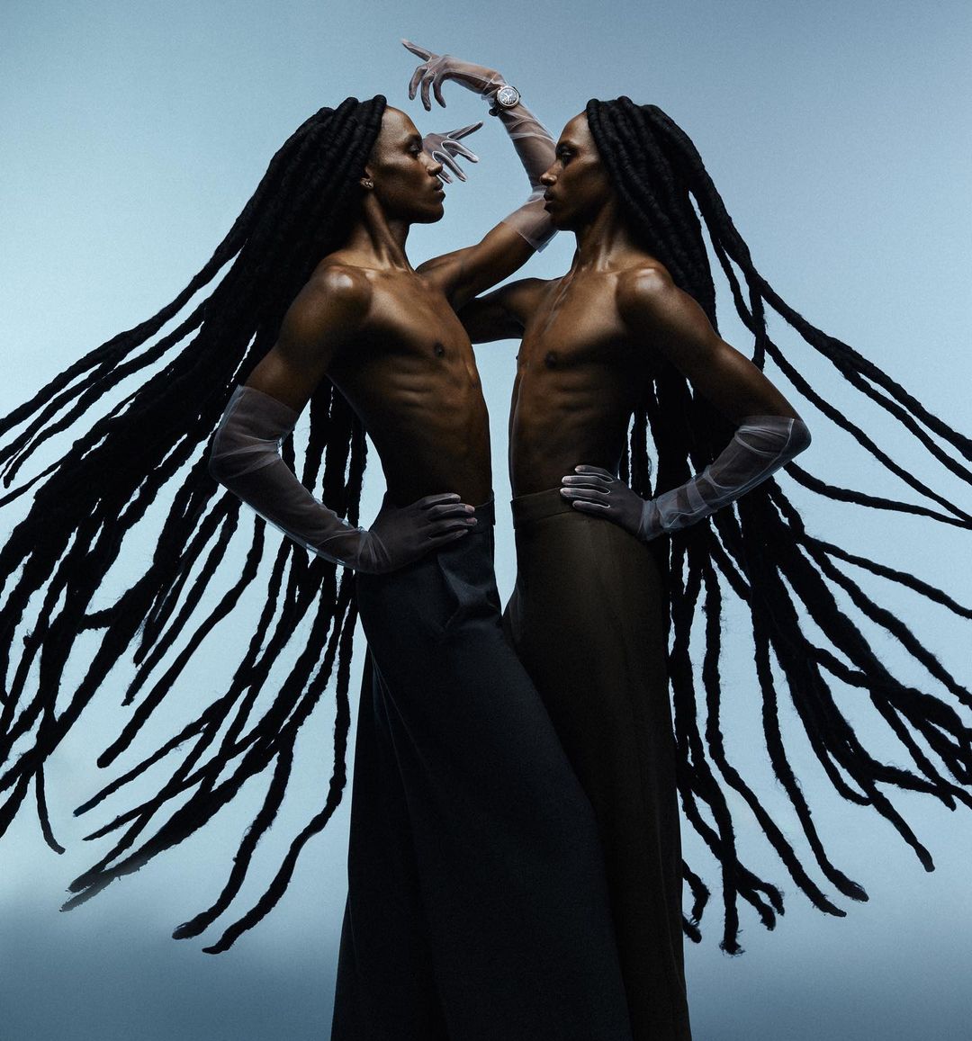 Two black men with long locs wearing sheer gloves and dark trousers posing against a blue studio background