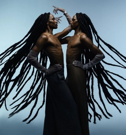 Two black men with long locs wearing sheer gloves and dark trousers posing against a blue studio background
