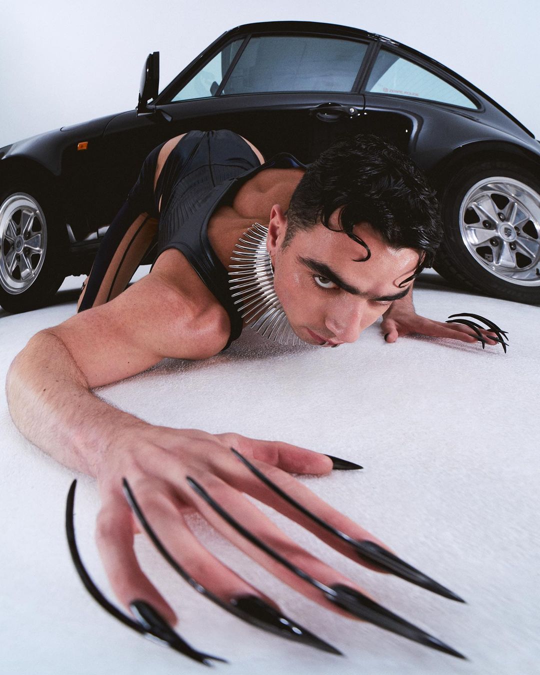 Adult male lies on a white surface with black stileto nails, wearing a spiked collar, in front of a black Porsche car in photo studio.