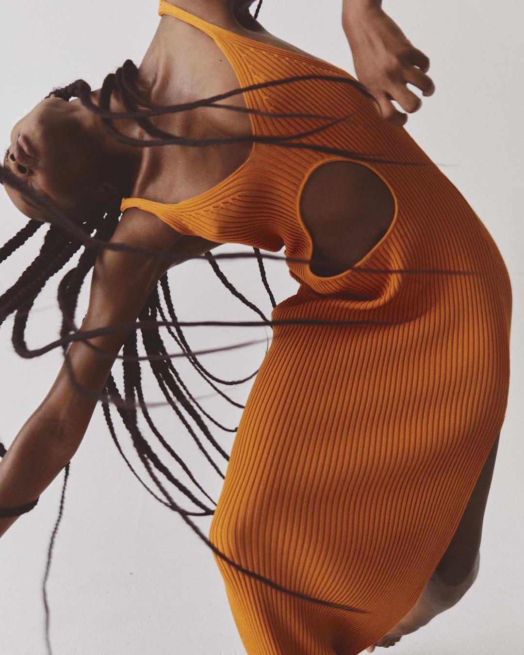 A Black woman with long braided hair in an orange knit dress poses against a white backdrop in a studio. Fashion editorial shot.