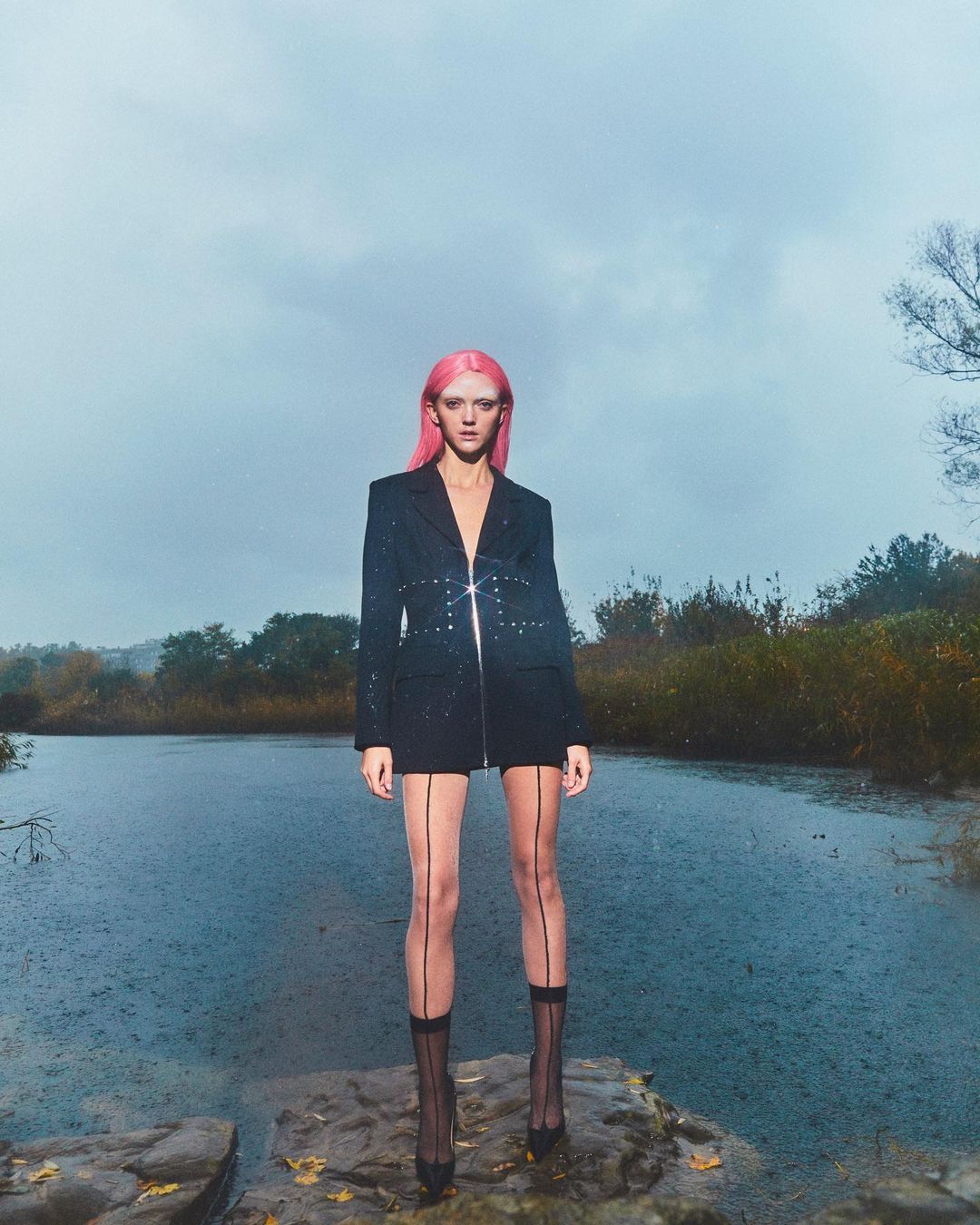 A stylish woman with pink hair, wearing a blazer dress with sheer stockings, poses near a reflective pool in a moody outdoor setting.