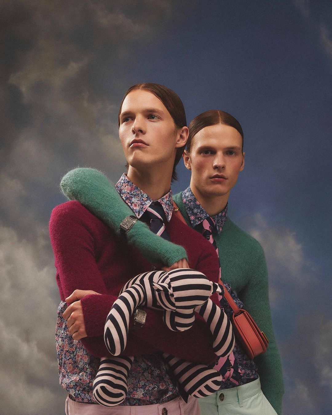 Two men in floral shirts and sweaters pose with a striped stuffed animal against a cloudy sky background in a fashion editorial shot.