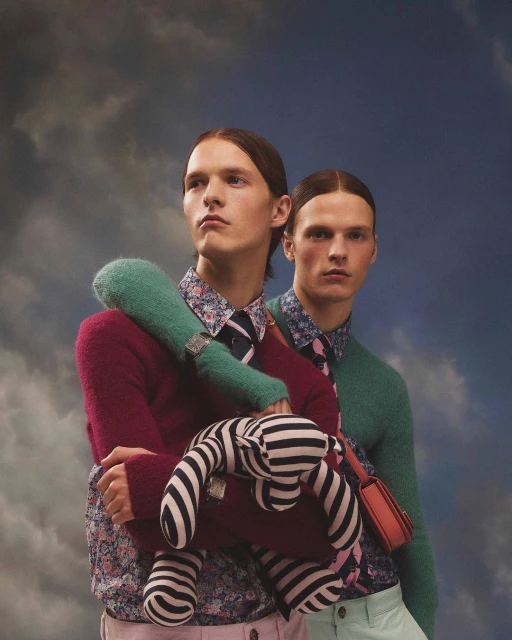 Two men in floral shirts and sweaters pose with a striped stuffed animal against a cloudy sky background in a fashion editorial shot.