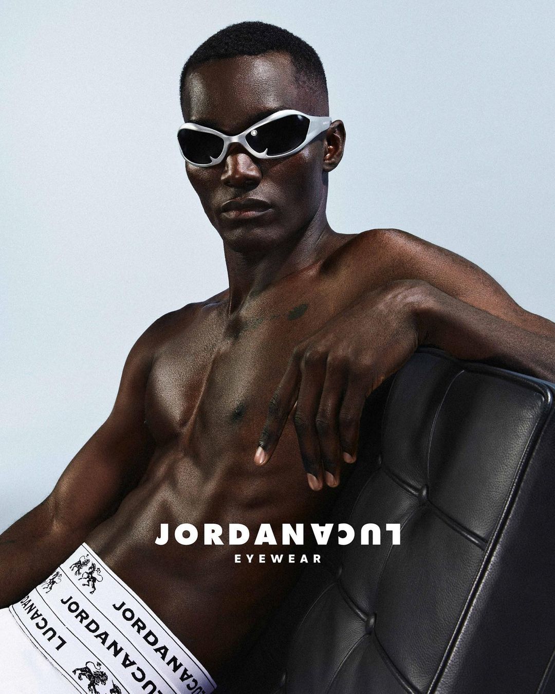 A bare-chested Black male model with silver sunglasses poses with a black tufted leather chair in a fashion-style studio shot.