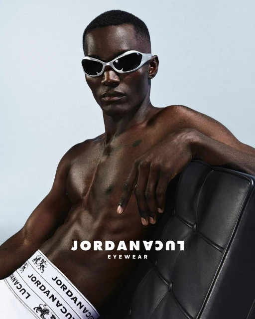 A bare-chested Black male model with silver sunglasses poses with a black tufted leather chair in a fashion-style studio shot.