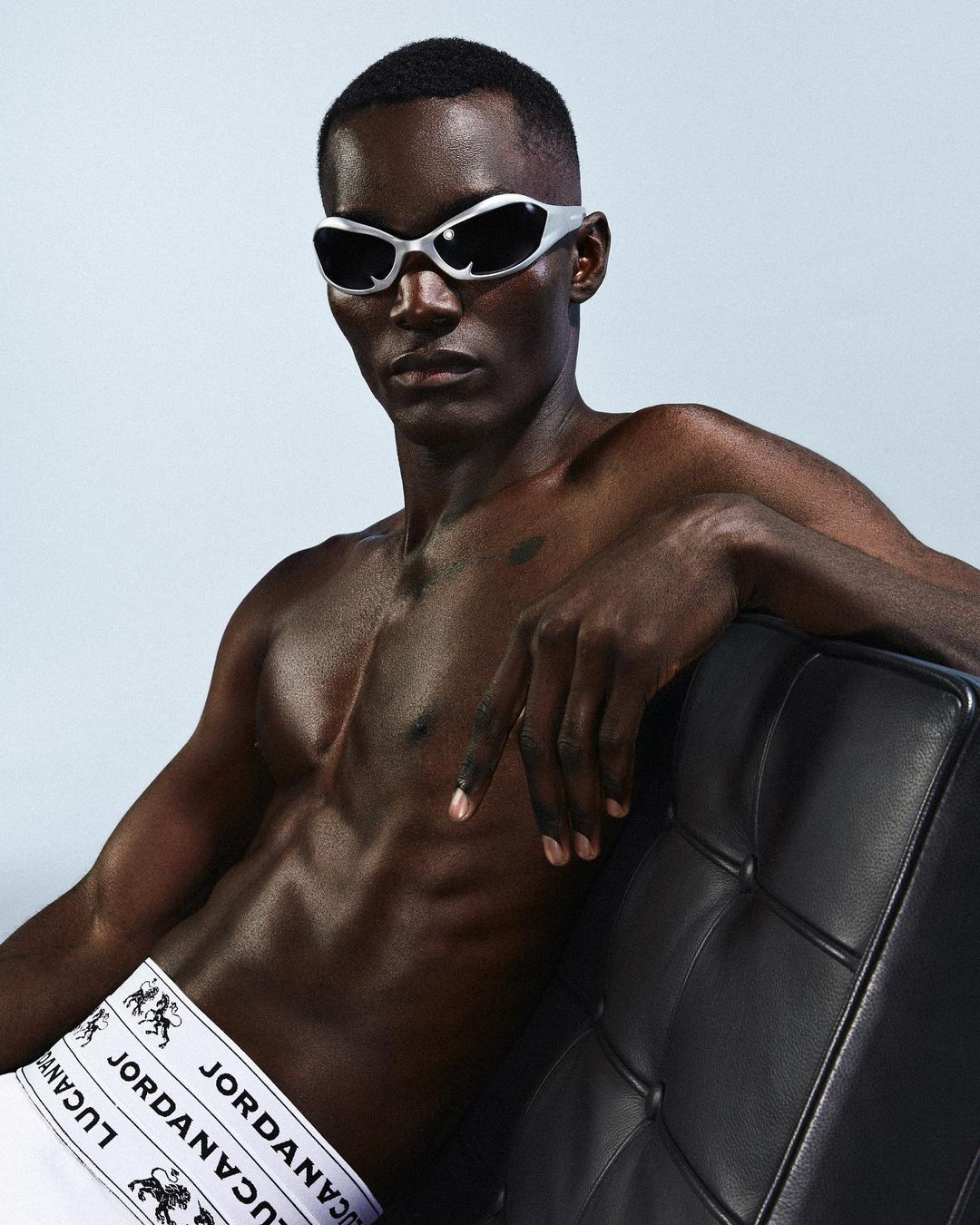 Athletic African male model poses shirtless, wearing silver sunglasses and underwear with text logo against a gradient grey background.