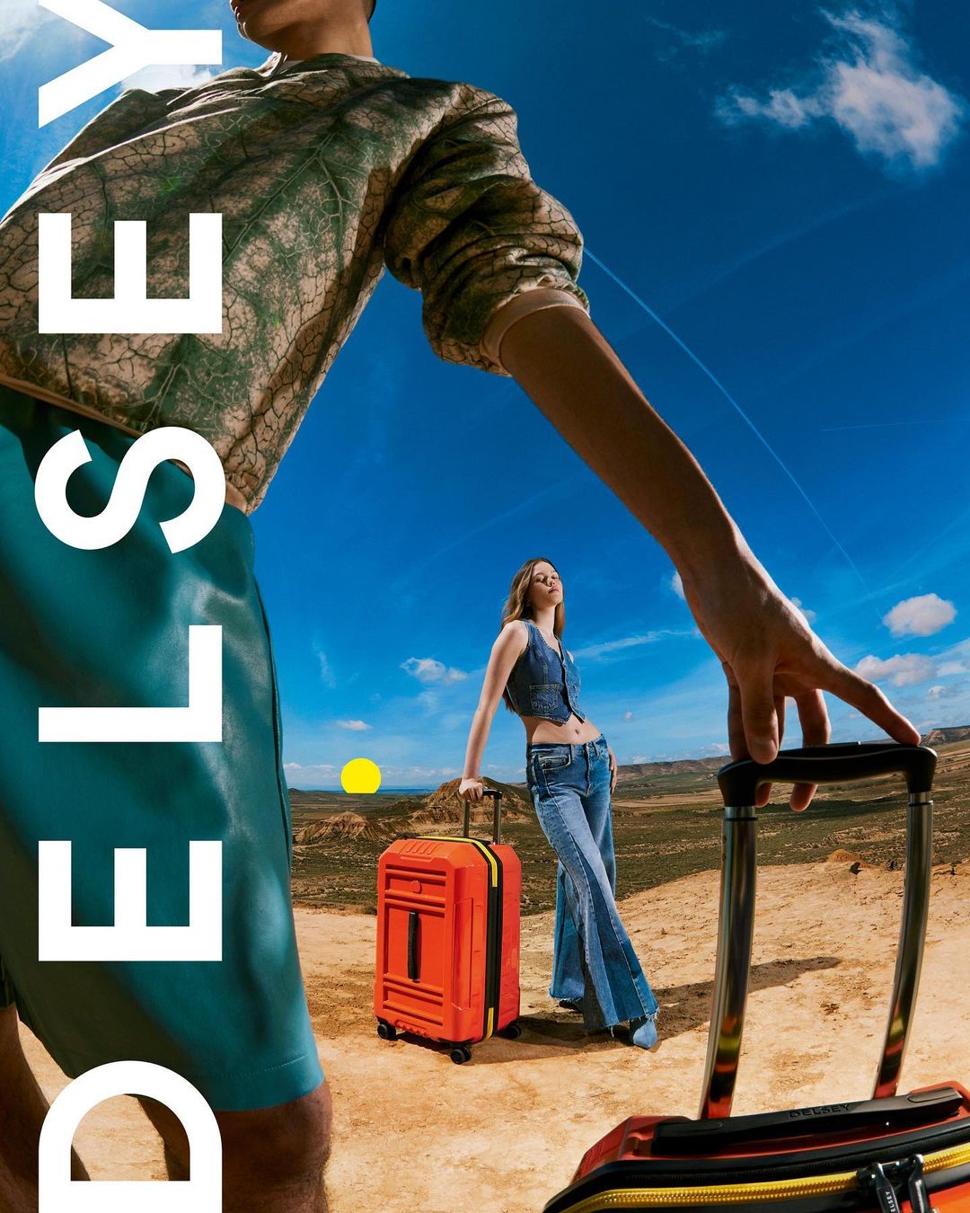 Two fashion models pose with luggage in a desert landscape under a blue sky, showcasing a travel-themed editorial campaign.