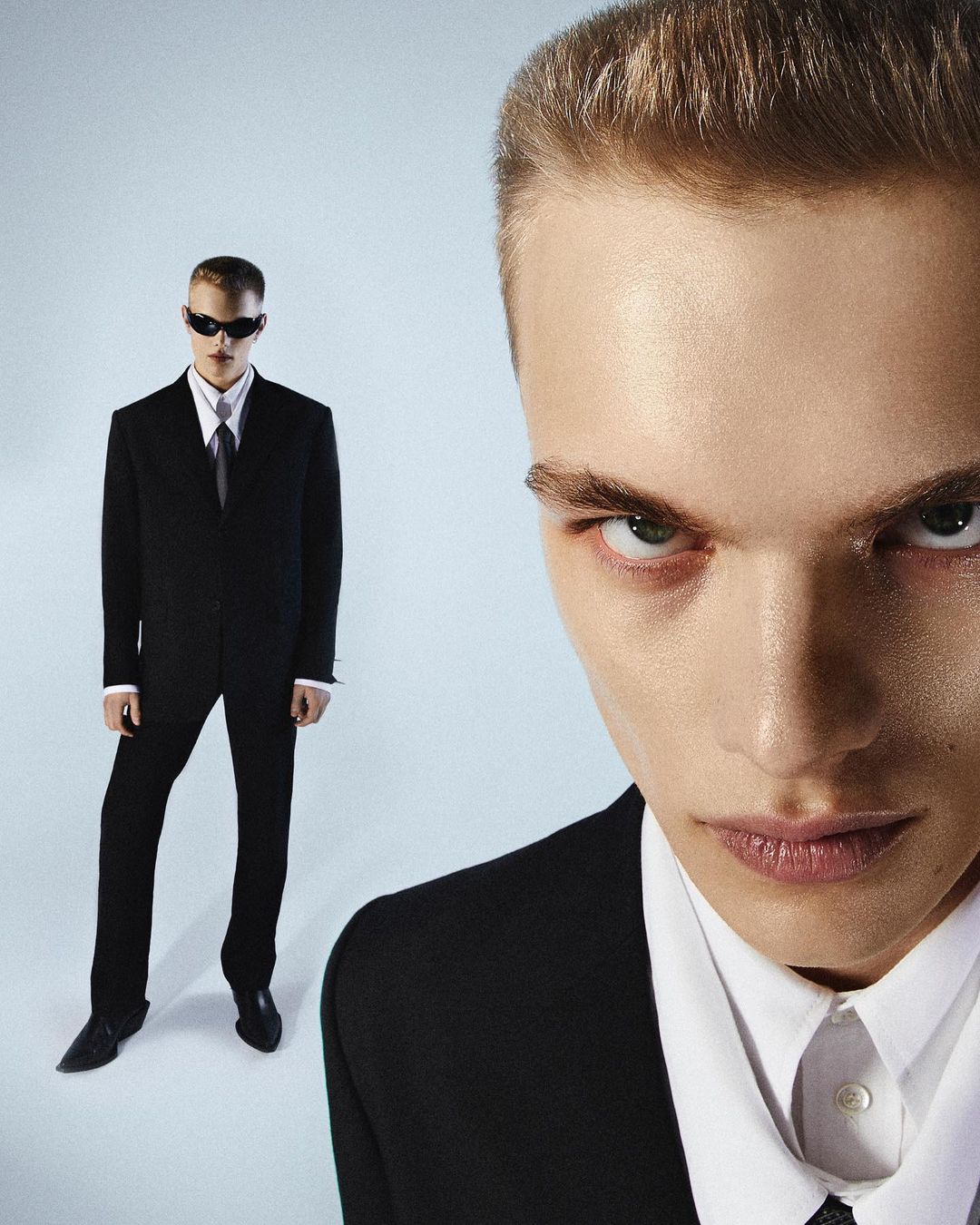Extreme close-up portrait of a man in a black suit with a second full-length figure standing in the background against a light backdrop
