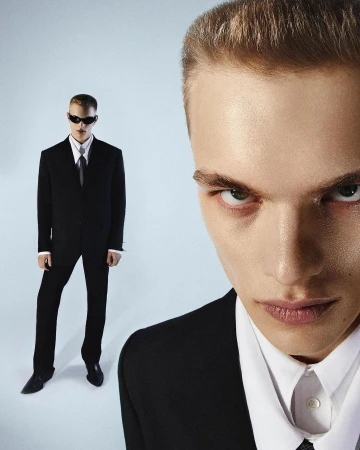 Extreme close-up portrait of a man in a black suit with a second full-length figure standing in the background against a light backdrop