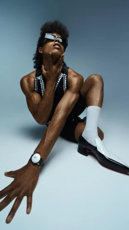 Person with afro and sunglasses poses in a sleeveless shirt, black shorts, white socks, and dress shoes against a light blue background.