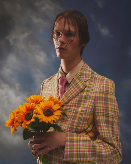 Stylish person in a plaid blazer holds yellow sunflowers against a cloudy sky. Fashion-forward portrait, unique style, eye-level framing.