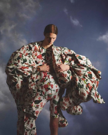 A person in a floral print outfit poses against a cloudy blue sky, creating a surreal and fashionable visual. The outfit is oversized.