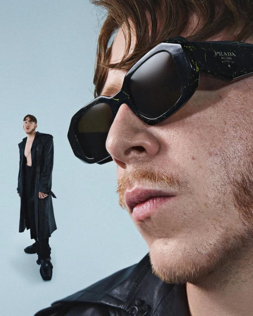 Close-up of a fair-skinned adult male with short brown hair and a mustache, wearing black Prada sunglasses and a black leather coat.
