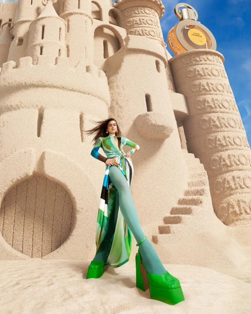 Woman posing in front of a large sandcastle with perfume bottle on top, wearing a green patterned dress and platform shoes in a studio.