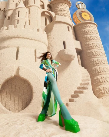 Woman posing in front of a large sandcastle with perfume bottle on top, wearing a green patterned dress and platform shoes in a studio.