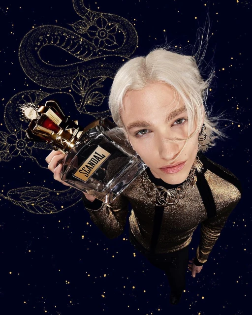 Female model with platinum hair holds Scandal perfume bottle against starry dark blue backdrop with gold snake illustration.