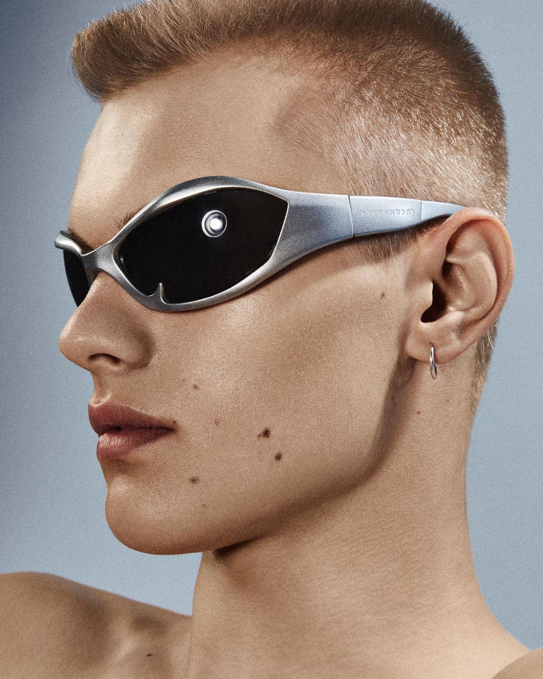 Portrait of a blonde male model wearing futuristic silver sunglasses against a light blue background in a studio setting.