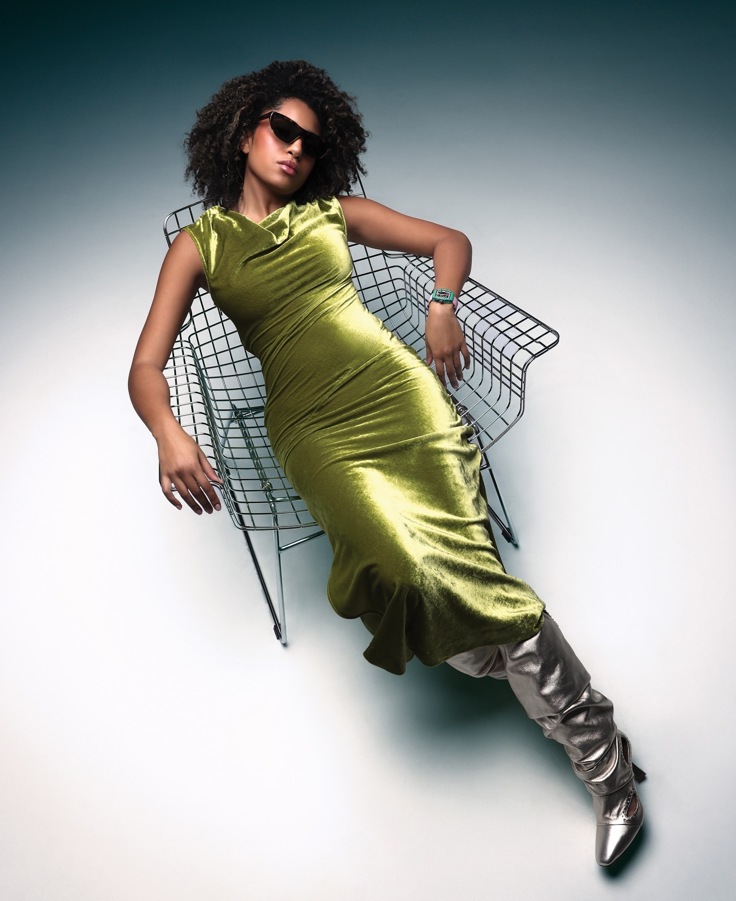 A woman with curly hair in sunglasses and a green velvet dress reclines in a wire chair, wearing silver thigh-high boots against a gradient background.