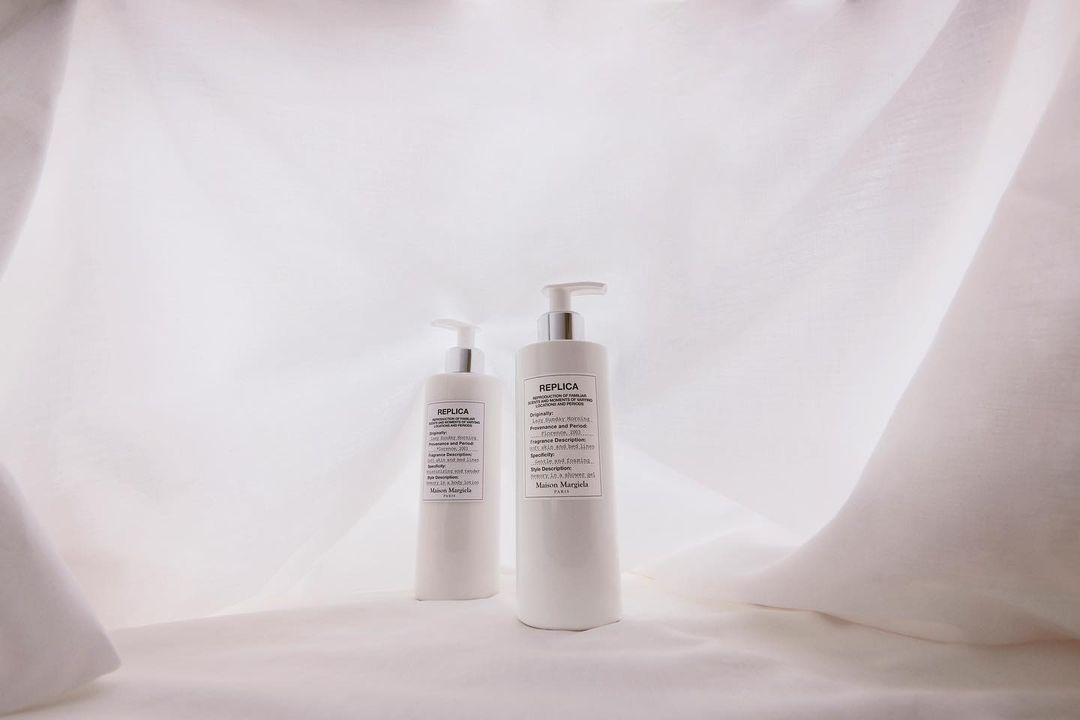 Two white bottles of Maison Margiela Replica fragrance stand on a white draped fabric background in soft, diffused lighting for a product shot.
