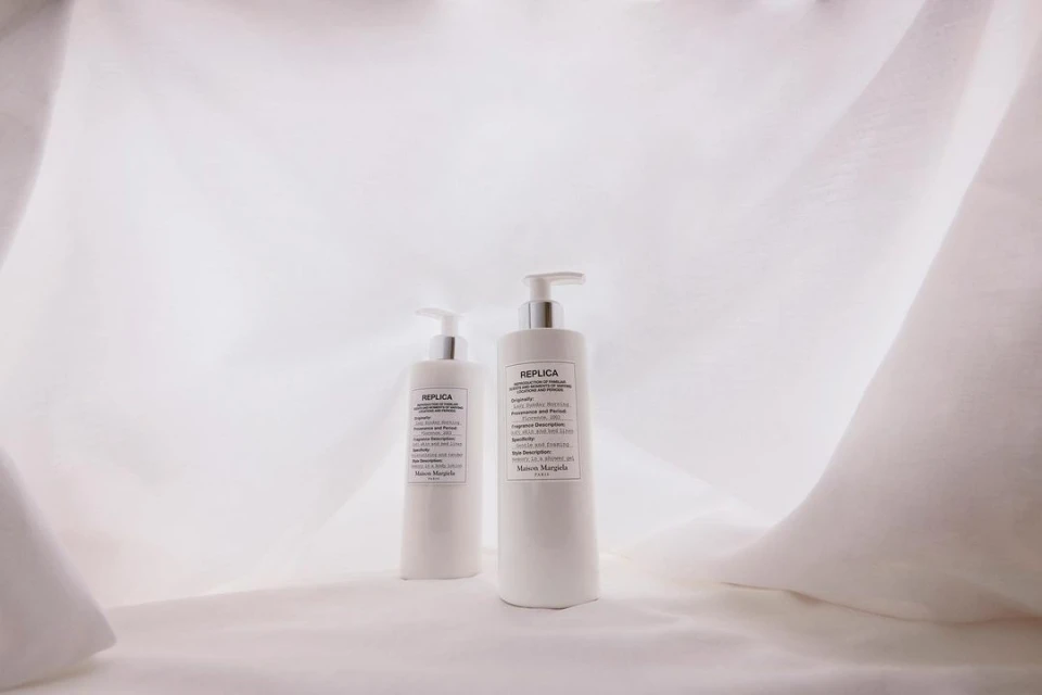 Two white bottles of Maison Margiela Replica fragrance stand on a white draped fabric background in soft, diffused lighting for a product shot.