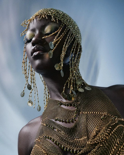 Woman with gold beaded headpiece and shoulder piece.  She has her eyes closed.  The background is light blue.