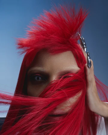 Portrait of a woman with bright red hair and long, silver-grey nails against a blue background in a beauty fashion shot.