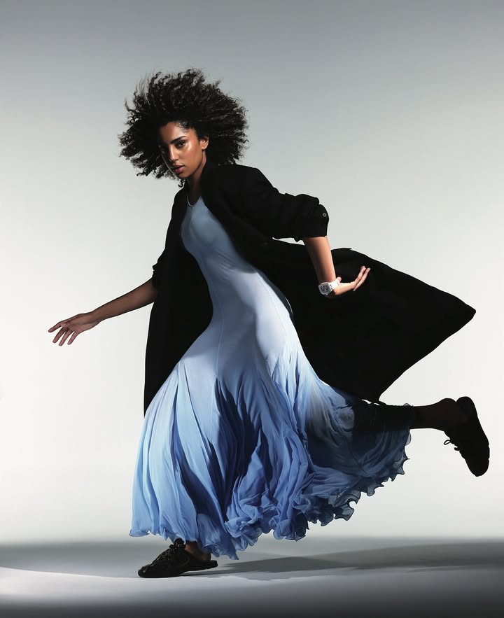 A woman with afro-style hair poses dynamically, wearing a light blue dress and dark coat in a studio setting, set against a gradient background.