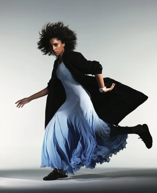A woman with afro-style hair poses dynamically, wearing a light blue dress and dark coat in a studio setting, set against a gradient background.