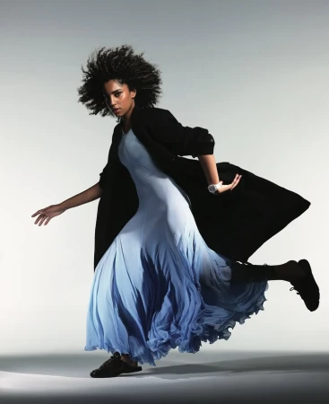 A woman with afro-style hair poses dynamically, wearing a light blue dress and dark coat in a studio setting, set against a gradient background.