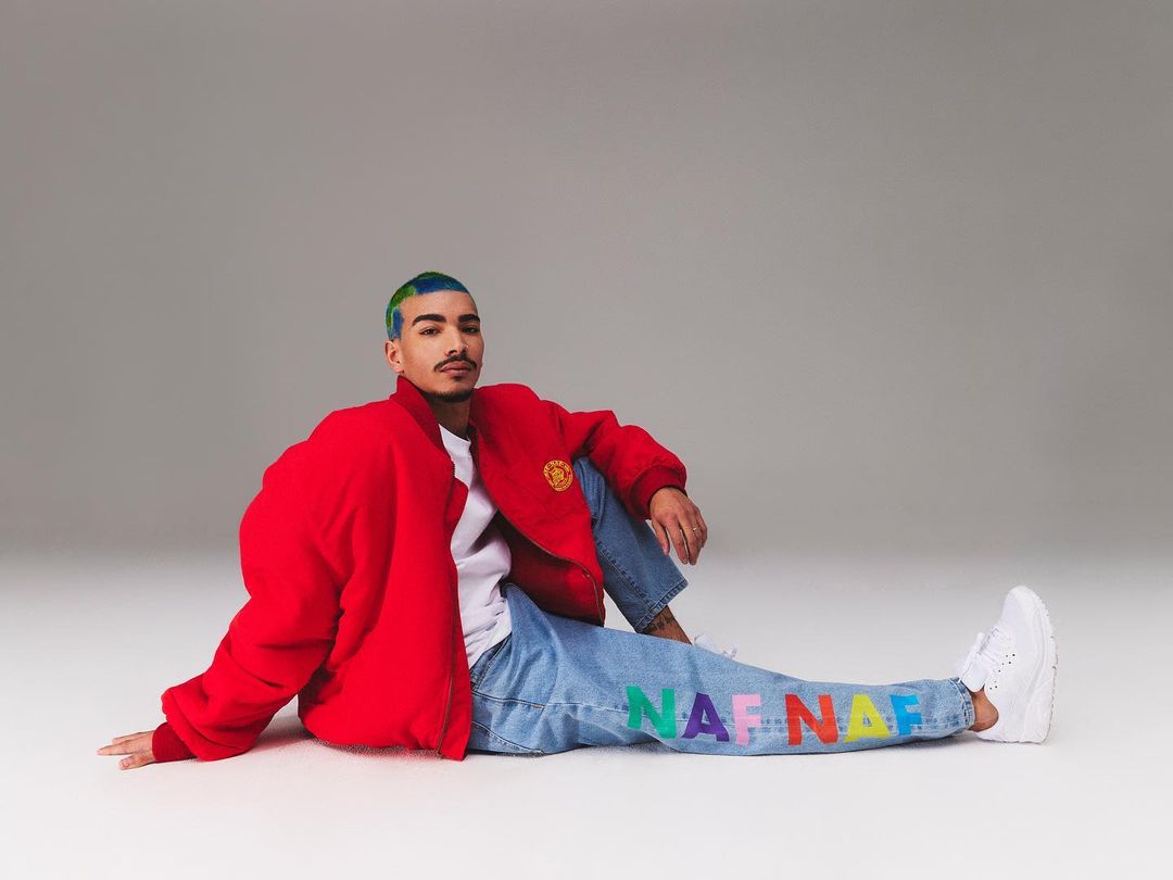 Man in red jacket sits on the floor in front of a gray backdrop wearing jeans and white sneakers. The jeans have colorful letters.