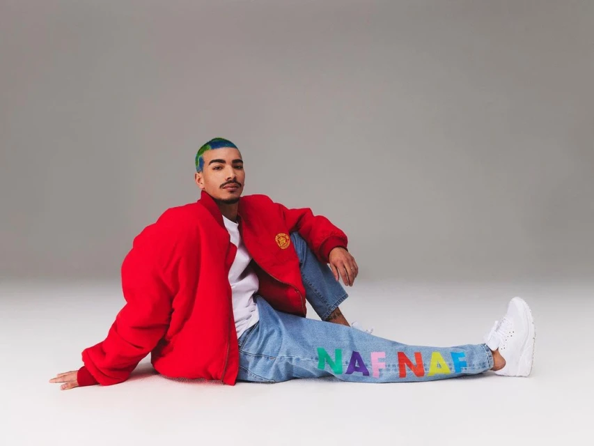 Man in red jacket sits on the floor in front of a gray backdrop wearing jeans and white sneakers. The jeans have colorful letters.