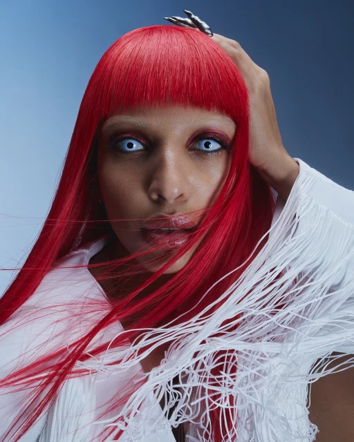 Portrait of a woman with red hair and white contact lenses wearing a white fringe top against a blue gradient background.