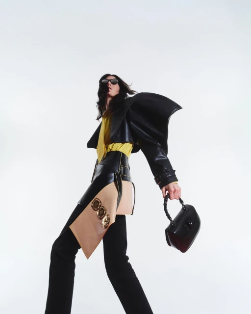 Fashionable adult strides in a studio, wearing a black leather jacket, yellow top, and avant-garde skirt, carrying a chic handbag.