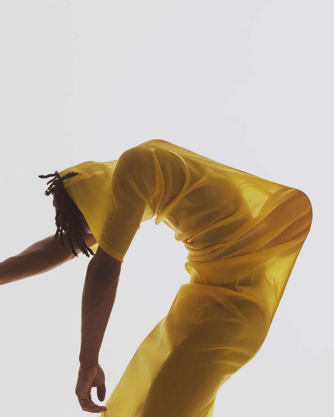 A person in a flowing yellow garment bends backward in a bright, minimalist studio setting showcasing fluid movement and fashion.