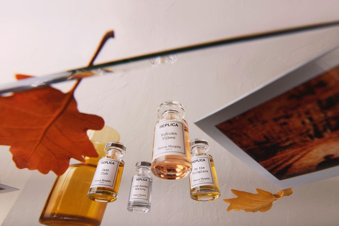 Overhead shot of assorted 'Replica' Maison Margiela fragrances with autumn leaves, arranged on a glass surface with a framed picture.