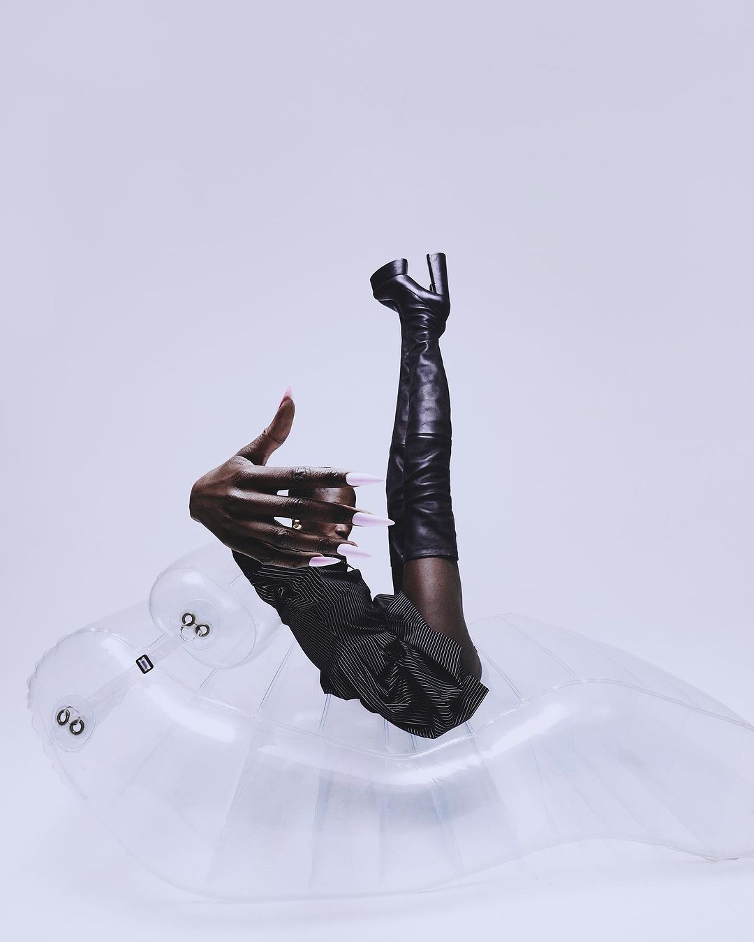 Fashion editorial featuring a dark-skinned person's hand with pink nails, striped garment, thigh-high leather boot, inflatable seat, and white backdrop.
