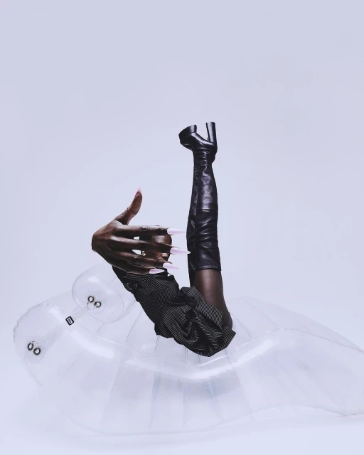 Fashion editorial featuring a dark-skinned person's hand with pink nails, striped garment, thigh-high leather boot, inflatable seat, and white backdrop.