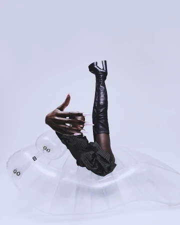 Fashion editorial featuring a dark-skinned person's hand with pink nails, striped garment, thigh-high leather boot, inflatable seat, and white backdrop.