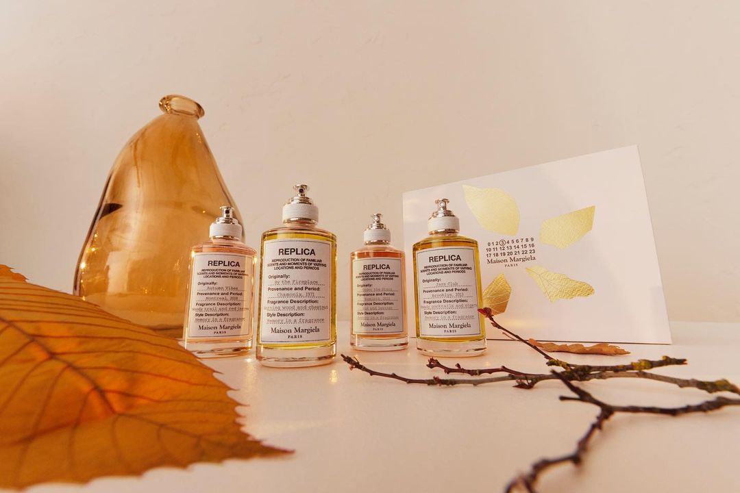 Four Maison Margiela REPLICA perfume bottles in amber tones arranged on a white surface, with a vase, leaves, and card as props.