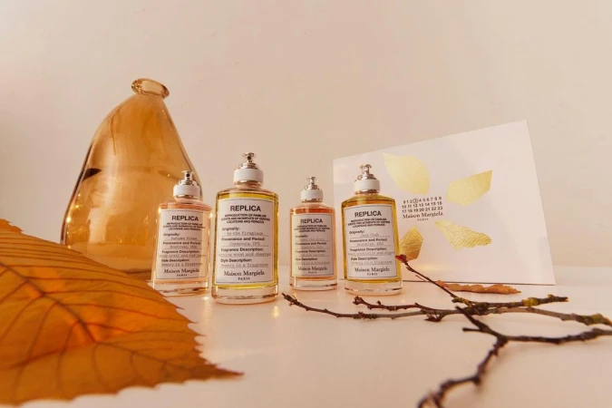 Four Maison Margiela REPLICA perfume bottles in amber tones arranged on a white surface, with a vase, leaves, and card as props.
