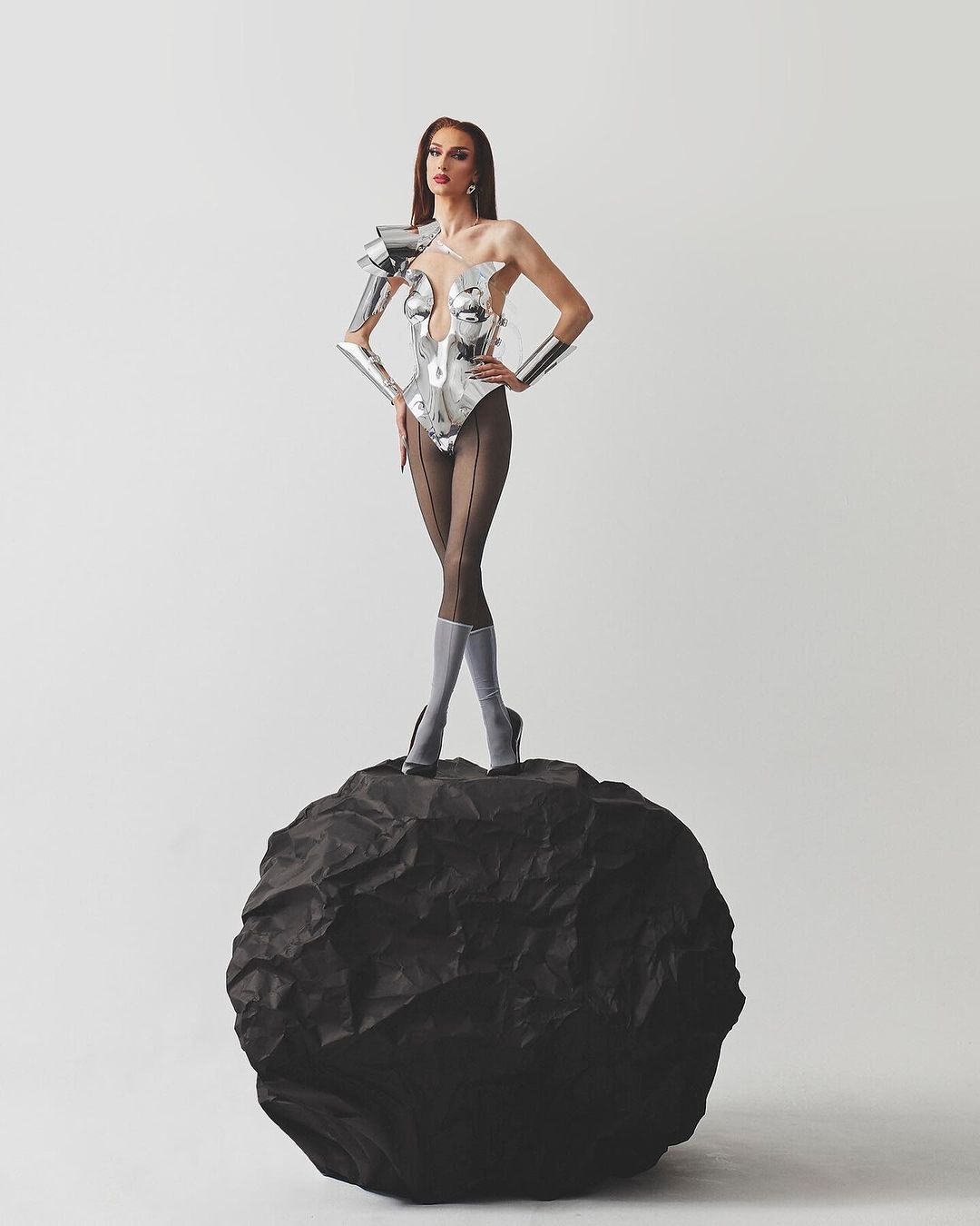 A slender woman with long brown hair, wearing a futuristic silver bodysuit and heels, poses confidently on a black rock in a studio setting.